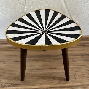 May include: A small three-legged table with a triangular top featuring a black and white sunburst pattern. The tabletop is enclosed by a thin gold-colored metal rim, and the three legs are made of dark brown wood with a smooth, tapered finish.