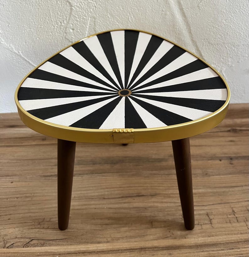 May include: A small three-legged table with a triangular top featuring a black and white sunburst pattern. The tabletop is enclosed by a thin gold-colored metal rim, and the three legs are made of dark brown wood with a smooth, tapered finish.