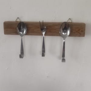 Könnte beinhalten: Ein hölzerner Utensilienhalter mit zwei Löffeln und einer Gabel aus Edelstahl. Der Halter ist an einer weißen Wand befestigt. Die Löffel und die Gabel sind an der Holzplatte befestigt.