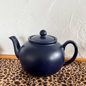 Könnte beinhalten: Eine marineblaue Keramik-Teekanne mit rundem Körper, gebogenem Ausguss und Schlaufengriff. Der Deckel hat einen kleinen, runden Knopf. Die Teekanne steht auf einer Oberfläche mit Leopardenmuster.
