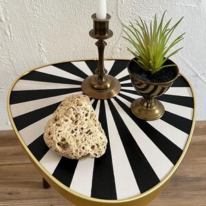 May include: A triangular side table with a black and white sunburst pattern on the top, featuring a gold-colored metal rim and dark wooden legs. On the table sits a brass candlestick with a white candle, a small green plant, and a porous rock.