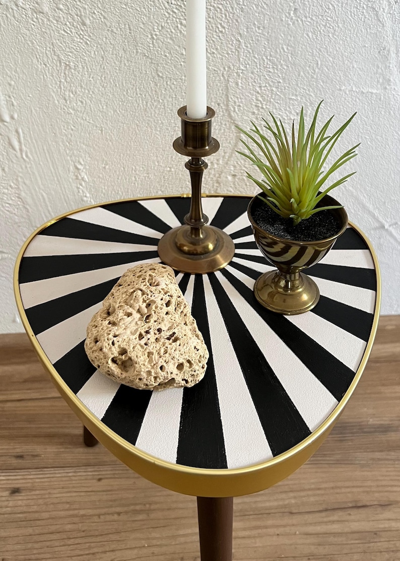 May include: A triangular side table with a black and white sunburst pattern on the top, featuring a gold-colored metal rim and dark wooden legs. On the table sits a brass candlestick with a white candle, a small green plant, and a porous rock.