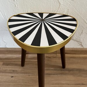May include: A three-legged side table featuring a triangular top with a black and white radial sunburst pattern. The tabletop is encased in a smooth gold-toned metal rim, and the three tapered legs are constructed from dark brown wood.