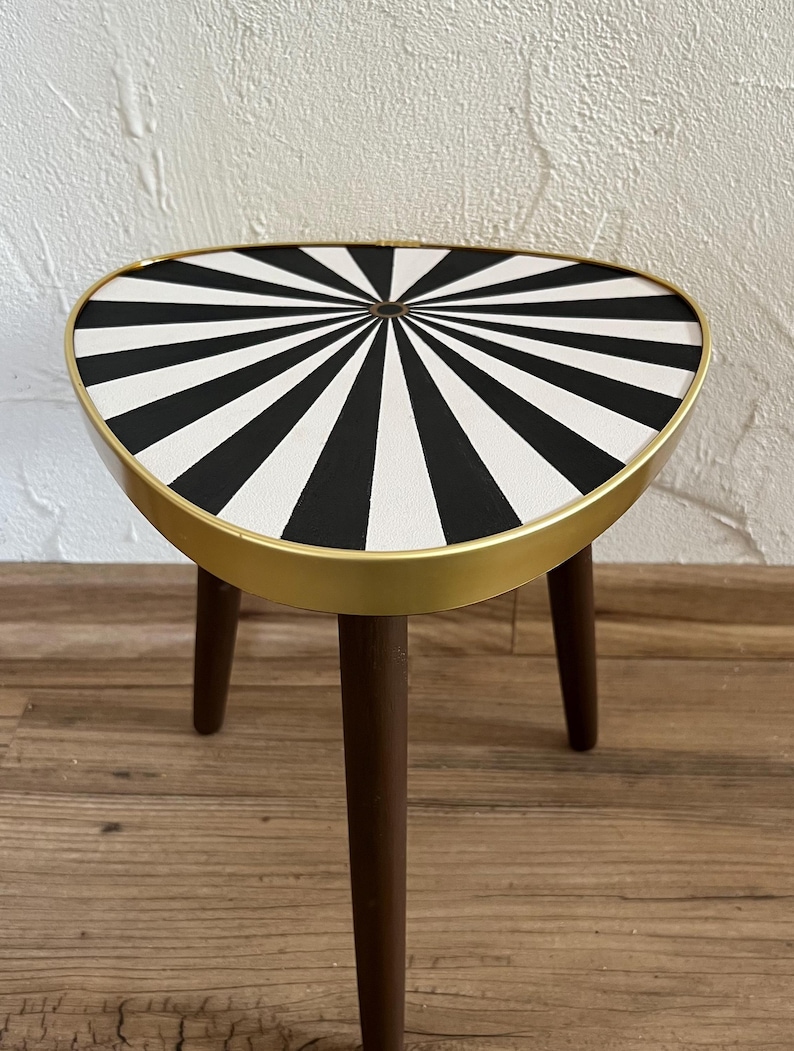 May include: A three-legged side table featuring a triangular top with a black and white radial sunburst pattern. The tabletop is encased in a smooth gold-toned metal rim, and the three tapered legs are constructed from dark brown wood.