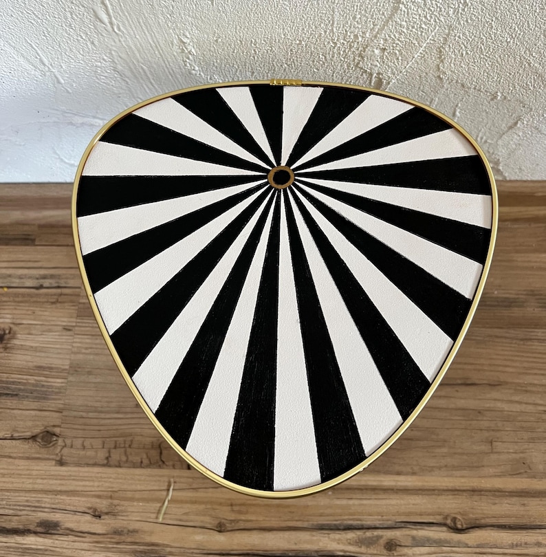 May include: A triangular side table with a rounded edge featuring a black and white sunburst pattern radiating from a central gold-rimmed circle. The tabletop is enclosed by a thin gold-colored metal frame that follows the curved triangular shape.