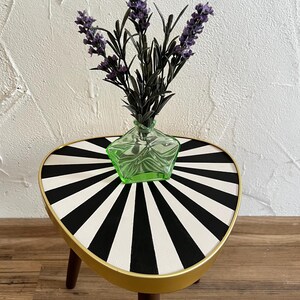 May include: A three-legged side table with a triangular top featuring a black and white sunburst pattern and a gold-toned rim. A small, star-shaped light green glass vase containing a sprig of purple lavender sits on the table surface.