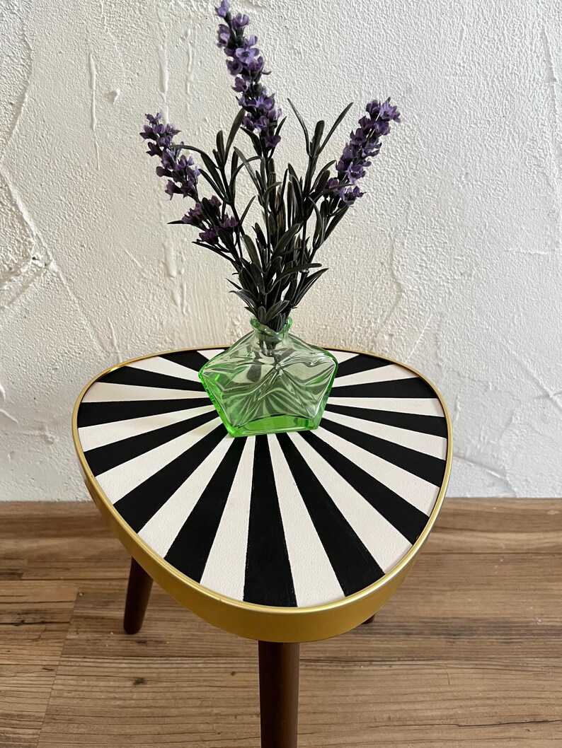 May include: A three-legged side table with a triangular top featuring a black and white sunburst pattern and a gold-toned rim. A small, star-shaped light green glass vase containing a sprig of purple lavender sits on the table surface.