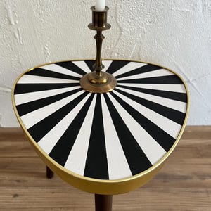 May include: A triangular side table with a black and white sunburst pattern on the top surface, a gold-toned metal rim, and dark wooden legs. A brass candlestick holder with a white taper candle is positioned in the center of the tabletop.
