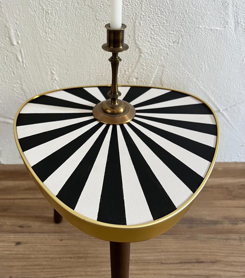 May include: A triangular side table with a black and white sunburst pattern on the top surface, a gold-toned metal rim, and dark wooden legs. A brass candlestick holder with a white taper candle is positioned in the center of the tabletop.