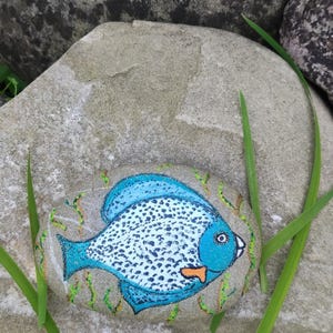 Könnte beinhalten: Ein handbemalter Stein mit einem blauen Fischdesign, weißen Punkten und einem orangefarbenen Maul. Der Fisch ist vor einem neutralfarbenen Stein platziert, mit grünen Wasserpflanzendetails. Das Kunstwerk ist von grünen Grashalmen umgeben.