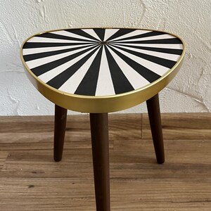 May include: A small three-legged side table featuring a triangular top with a black and white radial sunburst pattern. The tabletop is encased in a smooth gold-toned metal rim, and the three legs are made of dark brown wood with a tapered design.