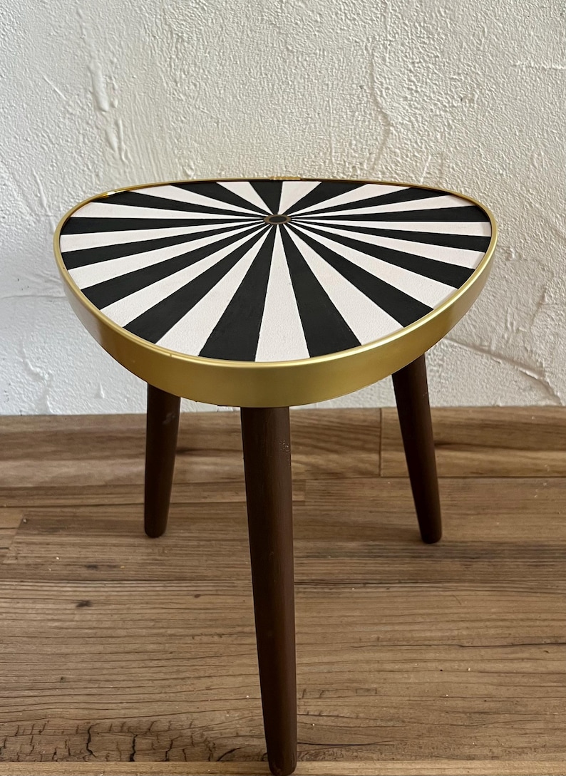 May include: A small three-legged side table featuring a triangular top with a black and white radial sunburst pattern. The tabletop is encased in a smooth gold-toned metal rim, and the three legs are made of dark brown wood with a tapered design.