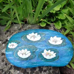 Könnte beinhalten: Ein handbemalter Stein, der einen blauen Teich mit fünf weißen Seerosen darstellt. Der Stein ist mit grünen Seerosenblättern und blauem Wasser mit weißen Wirbeln bemalt.