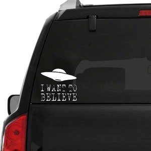 K&ouml;nnte beinhalten: Wei&szlig;er Vinyl-Aufkleber mit einem UFO und dem Text "I WANT TO BELIEVE". Der Aufkleber ist auf der Heckscheibe eines schwarzen Fahrzeugs angebracht. Das UFO hat die klassische Form einer fliegenden Untertasse.
