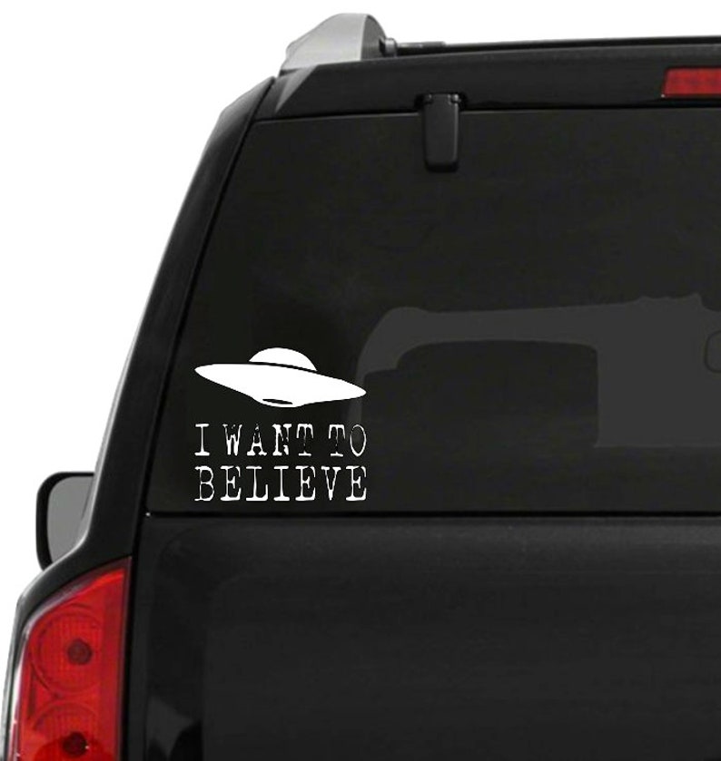 K&ouml;nnte beinhalten: Wei&szlig;er Vinyl-Aufkleber mit einem UFO und dem Text "I WANT TO BELIEVE". Der Aufkleber ist auf der Heckscheibe eines schwarzen Fahrzeugs angebracht. Das UFO hat die klassische Form einer fliegenden Untertasse.