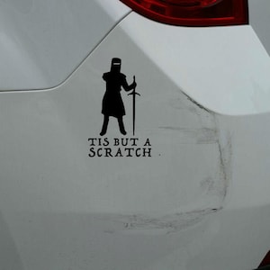 May include: Black vinyl decal of a knight holding a sword, with the text "TIS BUT A SCRATCH" below. The decal is applied to a white car, with visible scratches on the car's surface.