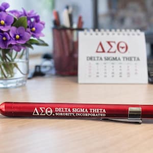 May include: A red pen with the Delta Sigma Theta Sorority, Incorporated logo and text. A calendar with the sorority's Greek letters and name is in the background, along with a vase of purple flowers.