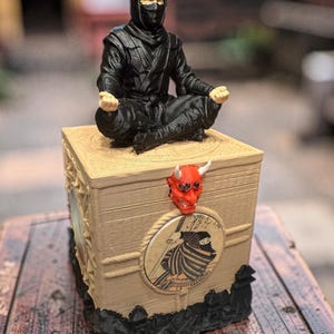 Op de afbeelding: Een zwarte ninja-figuur in een meditatieve pose zit op een beige doos, versierd met een rood demonenmasker en een ronde ninja-illustratie. De doos heeft een touwontwerp en zwarte accenten.