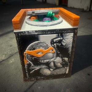 May include: A decorative box featuring the Teenage Mutant Ninja Turtles. The top has a manhole cover design with turtle faces. The sides display a black and white illustration of a turtle with an orange mask. The box has orange and white accents.