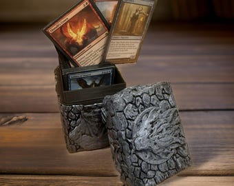 May include: A gray stone-textured box with a silver dragon design on the lid. The box is open and contains a stack of Magic: The Gathering cards, including "Thimma's Crown", "Bombard", and "Siege-Gang Commander".
