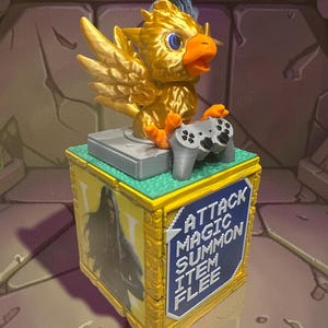 May include: A golden-yellow Chocobo bird figurine with blue eyes and an orange beak, holding a grey video game controller. The bird sits on a grey game console atop a yellow and blue box with the text "ATTACK MAGIC SUMMON ITEM FLEE".