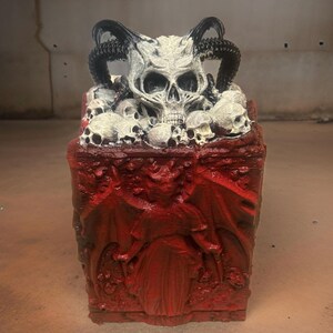 Puede incluir: Una caja roja con tem&aacute;tica demon&iacute;aca con un cr&aacute;neo y cuernos en la parte superior. La caja est&aacute; decorada con un relieve de un demonio con alas. La caja est&aacute; hecha de un material duro y tiene un acabado liso.