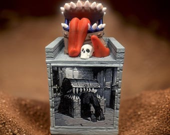 May include: A miniature dungeon trap with a painted scene of a chest with teeth and a skull on top. The trap is open and has a red tongue and teeth inside.