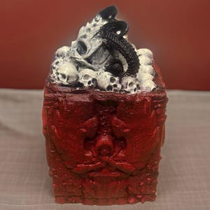 Puede incluir: Una caja roja, tallada intrincadamente, con una tapa llena de cr&aacute;neos blancos y un objeto negro, retorcido, parecido a un tent&aacute;culo. La caja tiene un dise&ntilde;o detallado y ornamentado.