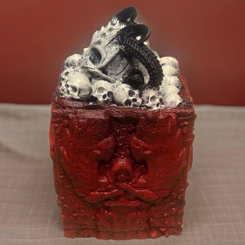 Puede incluir: Una caja roja, tallada intrincadamente, con una tapa llena de cr&aacute;neos blancos y un objeto negro, retorcido, parecido a un tent&aacute;culo. La caja tiene un dise&ntilde;o detallado y ornamentado.