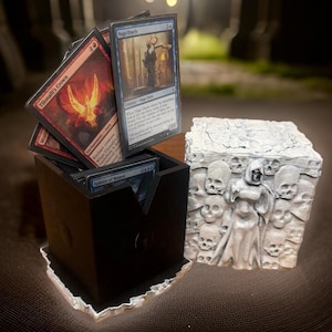 May include: A black and white 3D printed box with a skull and bone design. The box is open and has a stack of Magic: The Gathering cards inside. The cards are all different colors and have different illustrations.
