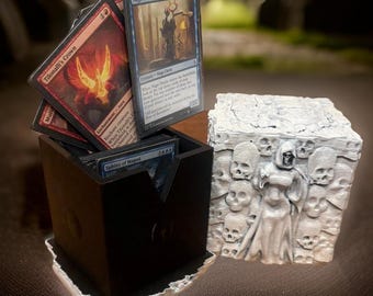 May include: A black and white 3D printed box with a skull and bone design. The box is open and has a stack of Magic: The Gathering cards inside. The cards are all different colors and have different illustrations.