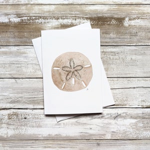 May include: A white card with a watercolor illustration of a sand dollar on a light brown wooden background.