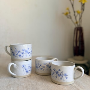 Pode incluir: Quatro canecas de cerâmica branca com um design floral azul. As canecas têm formato arredondado com alça e base natural sem esmalte. O padrão floral é em estilo delicado e desenhado à mão.