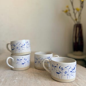 Pode incluir: Quatro canecas de cerâmica branca com desenhos florais azuis. Cada caneca tem uma base arredondada e uma alça curva. O padrão floral apresenta flores e caules azuis estilizados. As canecas estão empilhadas sobre uma superfície de madeira clara.