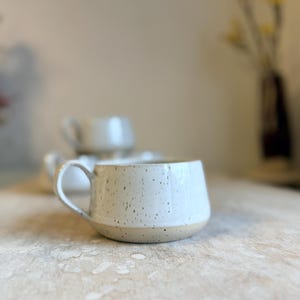 Può includere: Una tazza in ceramica bianca con un design maculato e una base arrotondata. La tazza ha un manico. Altre tazze sono visibili sullo sfondo. La tazza è su una superficie di legno.