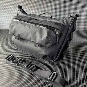 May include: Black messenger bag with a diamond pattern and multiple zippered compartments. Includes a detachable black strap with metal hardware. The bag has a top handle and a sleek, modern design.