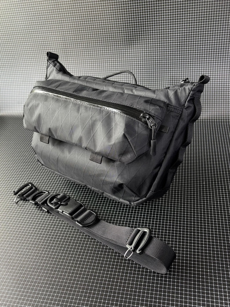 May include: Black messenger bag with a diamond pattern and multiple zippered compartments. Includes a detachable black strap with metal hardware. The bag has a top handle and a sleek, modern design.