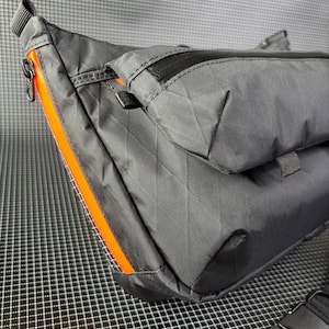 May include: A black nylon crossbody bag with a diamond pattern. The bag features an orange zipper and trim. The bag has a black adjustable strap and a black buckle. The bag has a small pocket on the front.