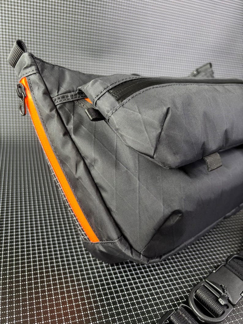 May include: A black nylon crossbody bag with a diamond pattern. The bag features an orange zipper and trim. The bag has a black adjustable strap and a black buckle. The bag has a small pocket on the front.