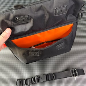 May include: Black messenger bag with an orange interior. The bag has a rectangular shape with a flap closure and a detachable black shoulder strap. The bag has a handle on top and a buckle closure on the strap.