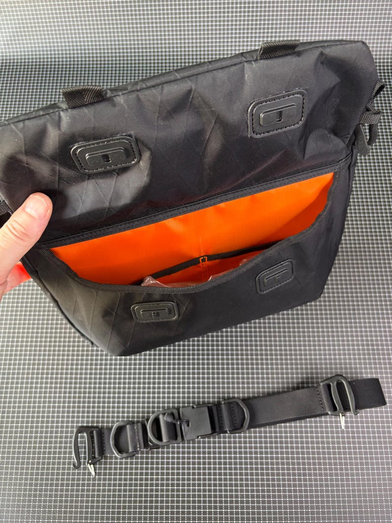 May include: Black messenger bag with an orange interior. The bag has a rectangular shape with a flap closure and a detachable black shoulder strap. The bag has a handle on top and a buckle closure on the strap.