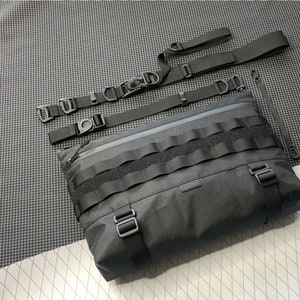 May include: A black messenger bag with a zippered closure and multiple adjustable straps. The bag has a large flap with a velcro closure and a series of black straps.