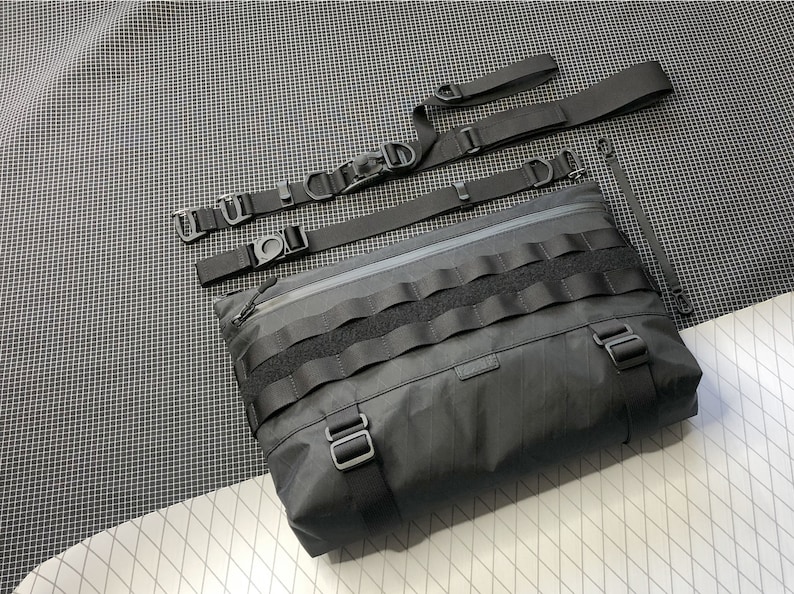 May include: A black messenger bag with a zippered closure, multiple straps, and a velcro flap with a row of velcro loops. Shoulder Bag, Messenger Bag, Techwear, X Pac, Sling Bag, EDC Bag, Commuter bag, lightweight bag, Cycling Bag, Laptop Bag, MacBook bag, D4bgs