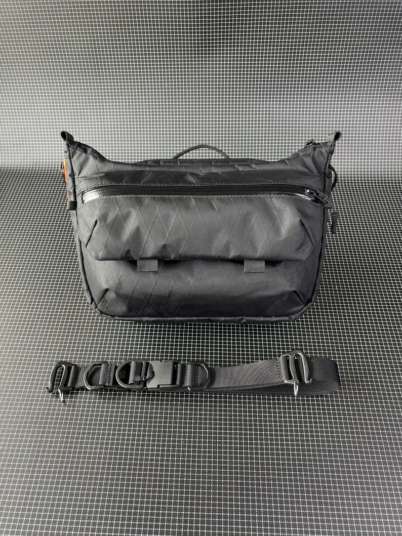 May include: A black messenger bag with a diamond pattern on the front. The bag has a top zipper, two front pockets, and a black strap with a buckle. The bag is designed for carrying items and has a handle on top.