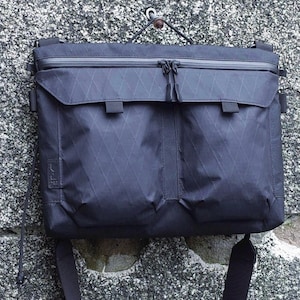 May include: A black messenger bag with a diamond pattern, hanging against a textured gray wall. The bag has a zippered top, two front pockets, and a shoulder strap. The bag is made of a durable, possibly waterproof material.