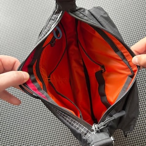 May include: Black crossbody bag with an orange interior. The bag has a zippered closure, a shoulder strap, and multiple interior pockets. The bag's exterior has a grid pattern.