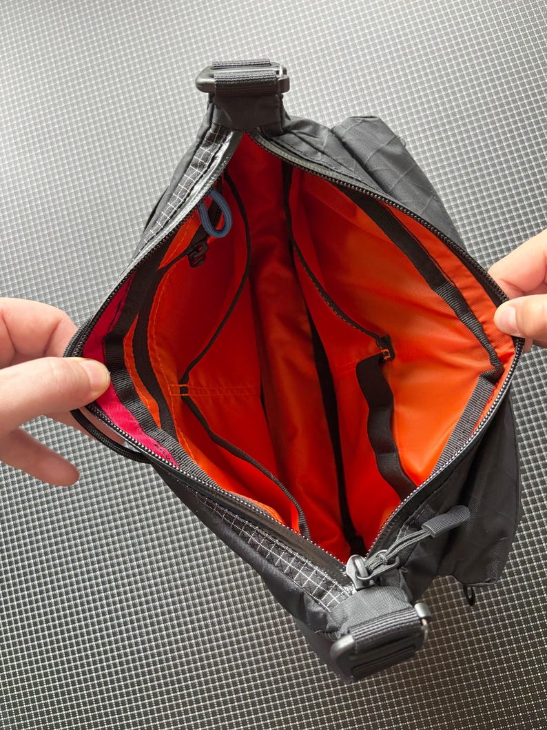 May include: Black crossbody bag with an orange interior. The bag has a zippered closure, a shoulder strap, and multiple interior pockets. The bag's exterior has a grid pattern.