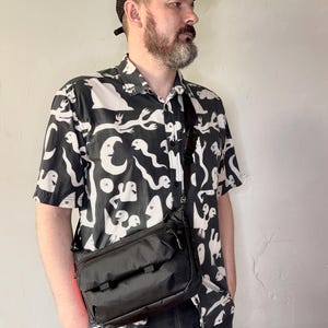May include: A black crossbody bag with a rectangular shape and multiple compartments. The bag is worn over the shoulder. The person is wearing a black and white patterned short-sleeve shirt and a black cap.