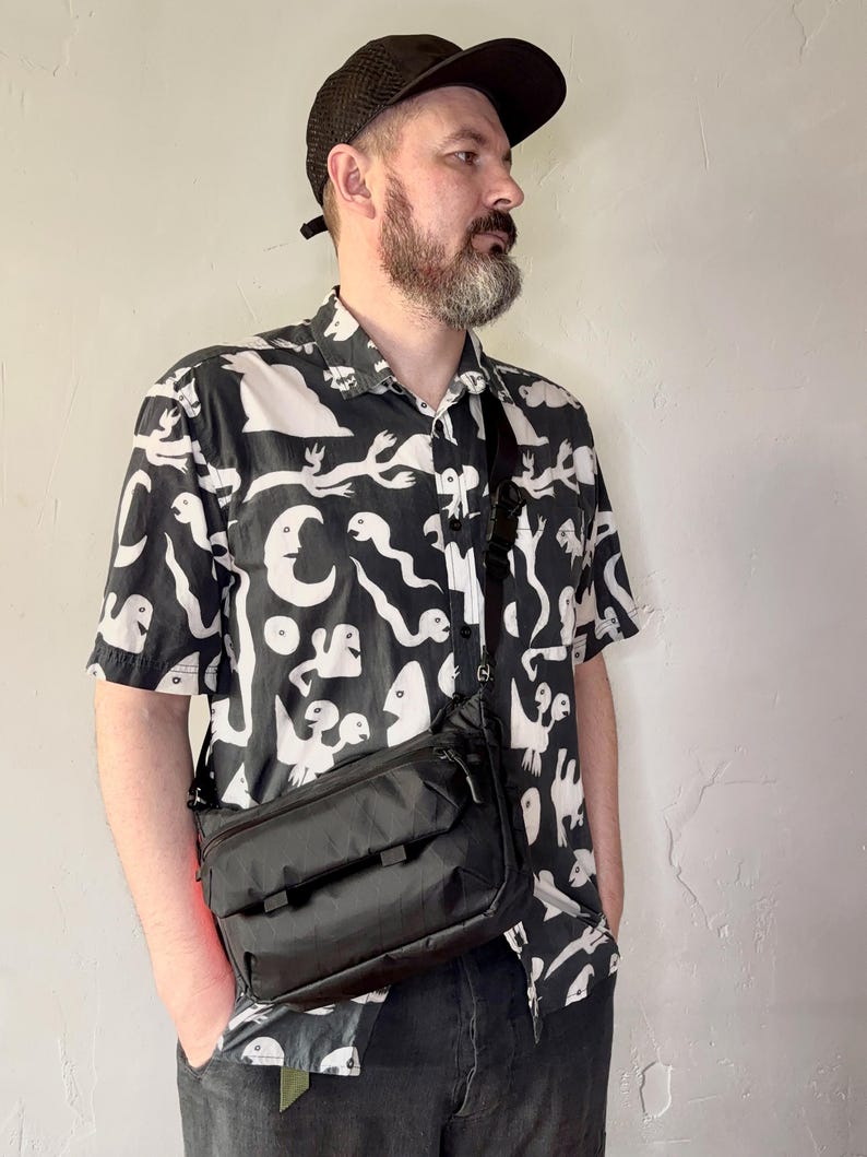 May include: A black crossbody bag with a rectangular shape and multiple compartments. The bag is worn over the shoulder. The person is wearing a black and white patterned short-sleeve shirt and a black cap.
