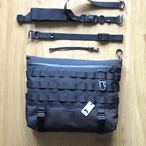 May include: A black messenger bag with a large front flap and multiple adjustable straps. The bag has a zipper closure and a small label with a logo on the front.